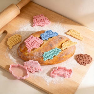 May include: A loaf of bread with cookie cutters spelling out "Hello" and the months of the year. The cutters are in pink, blue, yellow, and brown. A wooden rolling pin is in the background, with flour dusted around.