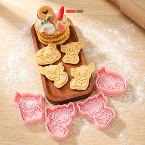 May include: A wooden tray displays baked cookies and pink cow-shaped cookie cutters. The cookies are light brown and decorated with a cow design. The pink cutters are in the shape of a cow. A rolling pin is in the background.