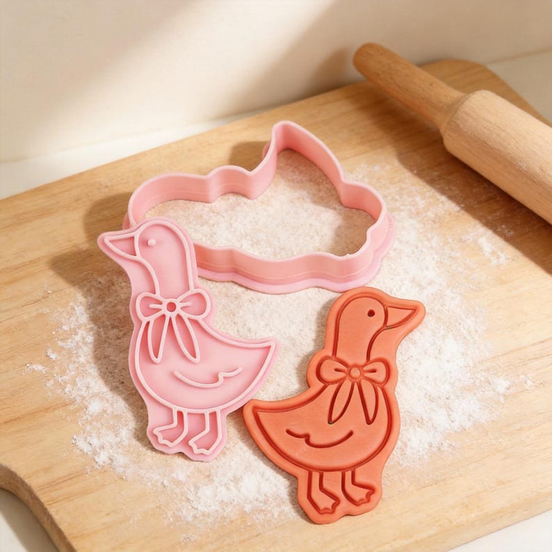 May include: Pink and orange goose-shaped cookie cutters with bow details, alongside a rolling pin, on a wooden cutting board dusted with flour. The cutters are in different shades of pink and orange.