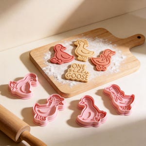 May include: A collection of goose-shaped cookie cutters and cookies. The cookies are in the shape of geese, some with bows and hats. The text "ONE SILLY GOOSE" is on one cookie. The cutters are pink, and the cookies are tan and red.