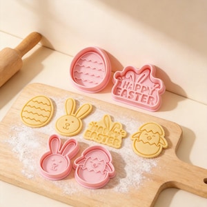 Könnte beinhalten: Eine Sammlung von Oster-Ausstechern in Rosa und Gelb. Das Set enthält Eier-, Hasen- und Kükenformen sowie einen Ausstecher mit der Aufschrift "Happy Easter". Die Ausstecher liegen auf einem Holzbrett.