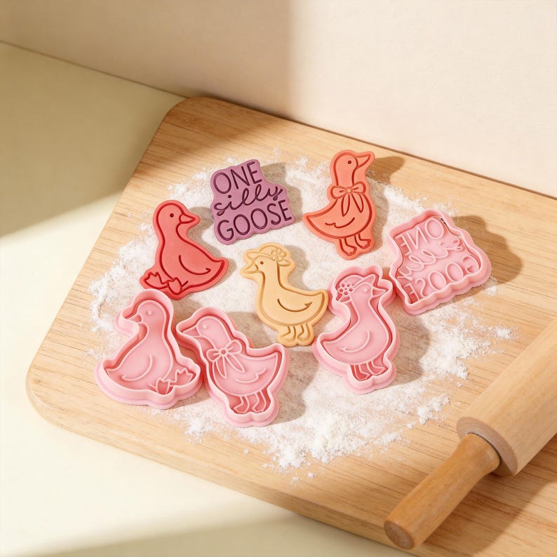 May include: A collection of colorful goose-shaped cookie cutters in shades of pink, orange, and yellow, arranged on a wooden cutting board dusted with flour. One cutter has the text "ONE silly GOOSE". A wooden rolling pin is in the background.