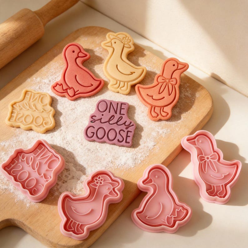 May include: A collection of goose-shaped cookie cutters and cookies in various colors, including pink, peach, and tan. Some cookies have a bow or hat design. One cookie has the text "ONE silly GOOSE". The items are arranged on a wooden cutting board with flour.