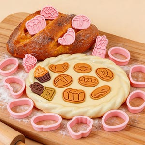 Puede incluir: Un conjunto de cortadores de galletas rosas en varias formas, incluyendo círculos y formas de pan, junto con una barra de pan y pasteles decorativos. Los cortadores están dispuestos sobre una superficie de madera, sugiriendo un tema de repostería.
