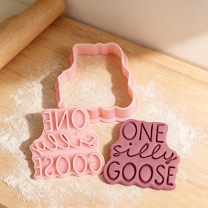 May include: Pink cookie cutters and stamp with the words "ONE silly GOOSE" on a wooden surface. The set includes a goose-shaped cutter and a stamp with the phrase in a playful font. A rolling pin is visible in the background.
