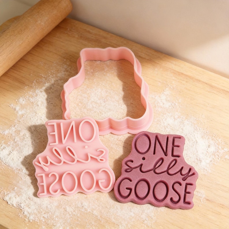 May include: Pink cookie cutters and stamp with the words "ONE silly GOOSE" on a wooden surface. The set includes a goose-shaped cutter and a stamp with the phrase in a playful font. A rolling pin is visible in the background.