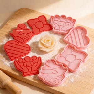 May include: A collection of red and pink cookie cutters in various shapes, including hearts, hands, and a family design, arranged on a wooden board with a white rose. The cutters are ideal for making cookies for special occasions.