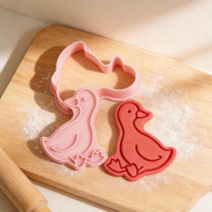 May include: Pink and red duck-shaped cookie cutters on a wooden cutting board dusted with flour. The cutters are in the shape of a duck, with details like a beak, eye, and webbed feet. A rolling pin is visible in the lower left corner.