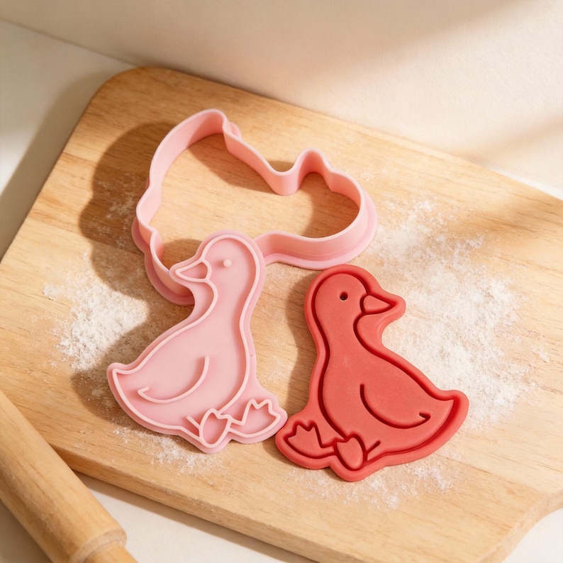 May include: Pink and red duck-shaped cookie cutters on a wooden cutting board dusted with flour. The cutters are in the shape of a duck, with details like a beak, eye, and webbed feet. A rolling pin is visible in the lower left corner.