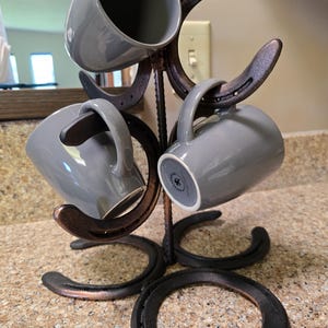 May include: A dark bronze horseshoe-shaped mug tree holds four grey ceramic mugs. The metal tree has horseshoes forming the mug holders. The mugs are a solid grey colour.