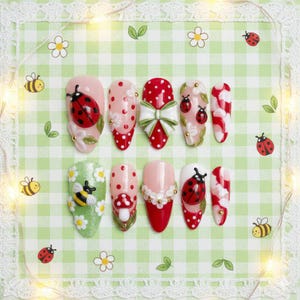 May include: A set of ten artificial nails with various designs. The nails feature ladybugs, flowers, polka dots, and a bow. Colors include red, white, pink, and green. Some nails have gold accents. The nails are arranged on a green and white checkered background.