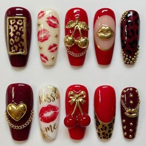 May include: A collection of ten artificial nails with various designs. The nails feature a range of colors, including red, white, and burgundy, and are adorned with gold accents, lip prints, and animal print patterns. Some nails have gold chains and heart or cherry designs.