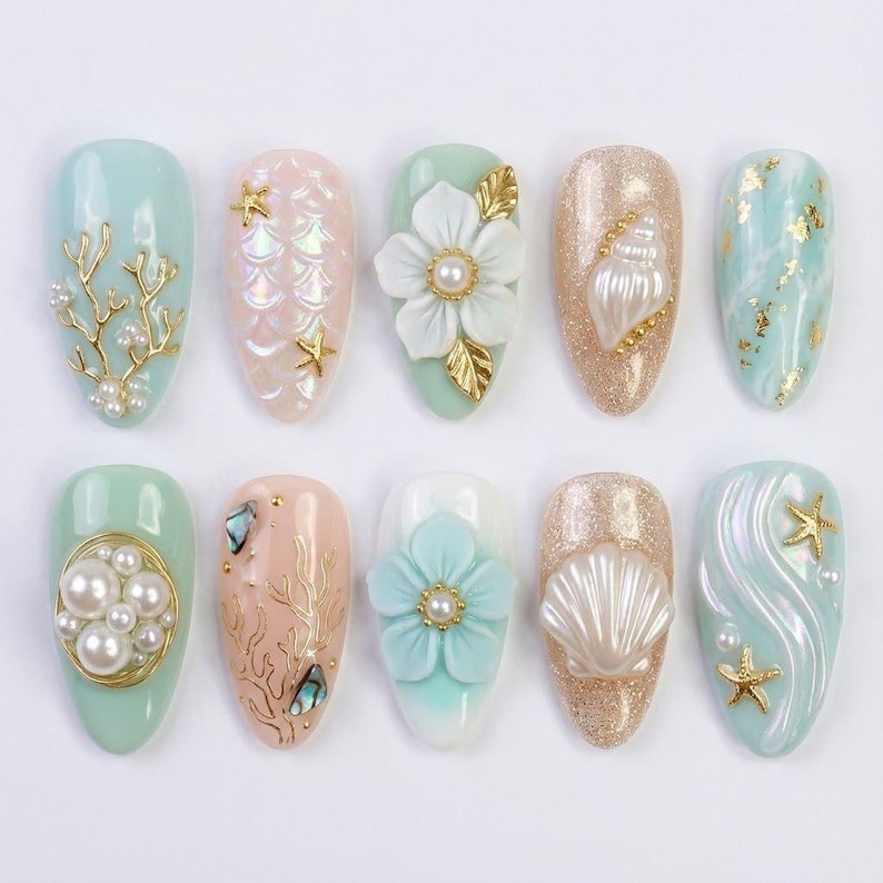 May include: A collection of ten artificial nails with various designs. The nails feature a range of colors, including light blue, pink, and gold, and are adorned with embellishments such as pearls, seashells, and floral accents. The designs evoke a marine theme.