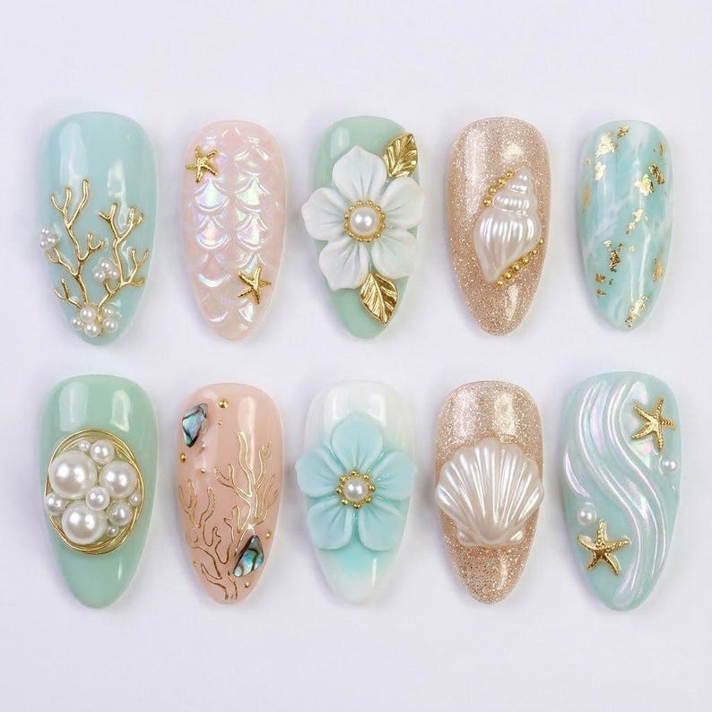 May include: A collection of ten artificial nails with various designs. The nails feature a range of colors, including light blue, pink, and gold, and are adorned with elements like pearls, seashells, and floral accents. The designs evoke a marine theme.