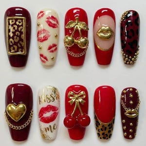 May include: A collection of ten artificial nails with various designs. The nails feature a range of colors, including red, white, and burgundy, and are adorned with gold accents, lip prints, and animal prints. Some nails have gold chains and charms.