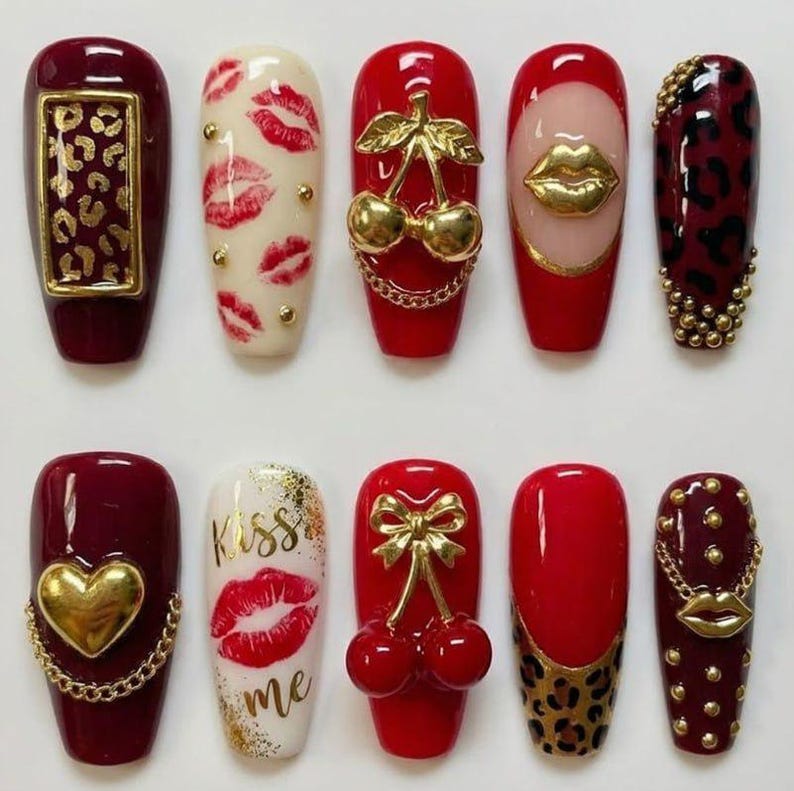 May include: A collection of ten artificial nails with various designs. The nails feature a range of colors, including red, white, and burgundy, and are adorned with gold accents, lip prints, and animal prints. Some nails have gold chains and charms.