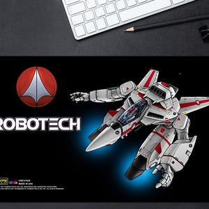 May include: A rectangular black desk mat featuring the white and red Robotech VF-1 Valkyrie mecha in flight mode. The design includes the red and white Robotech logo, the word ROBOTECH in white text, and small copyright information in the corner.