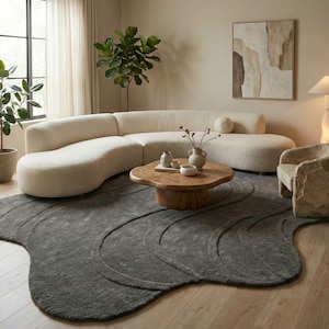 May include: A contemporary living room with a cream-coloured curved sofa, a dark grey rug with a wave design, and a wooden coffee table. The room is decorated with plants, an abstract painting, and a stone-like armchair.