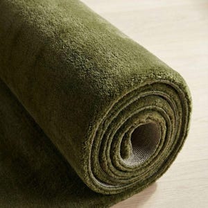May include: Rolled-up olive green rug on a light wood surface. The rug's plush texture is visible, with the rolled section showing the backing material. The color is a deep, earthy green.