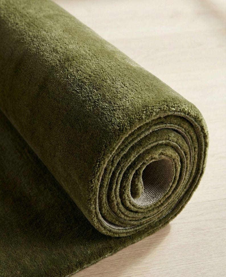 May include: Rolled-up olive green rug on a light wood surface. The rug's plush texture is visible, with the rolled section showing the backing material. The color is a deep, earthy green.