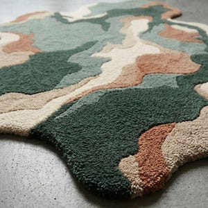 May include: An area rug with an irregular, organic shape featuring a tufted, high-pile texture. The design consists of abstract, flowing color blocks in shades of dark forest green, sage green, cream, and muted terracotta, creating a layered effect.