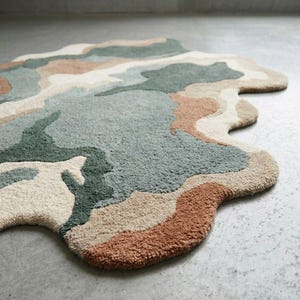 May include: An area rug with an irregular, organic shape featuring a tufted texture. The design consists of abstract, flowing patches of color in muted sage green, dark forest green, warm terracotta, cream, and beige tones across the surface.