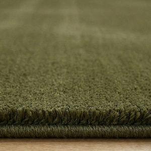 May include: Close-up of a plush, olive green rug. The rug has a soft, textured surface with a visible fringe along the edge. The color is a muted, earthy green, suggesting a natural or organic aesthetic. The rug appears to be made of a dense, high-pile material.