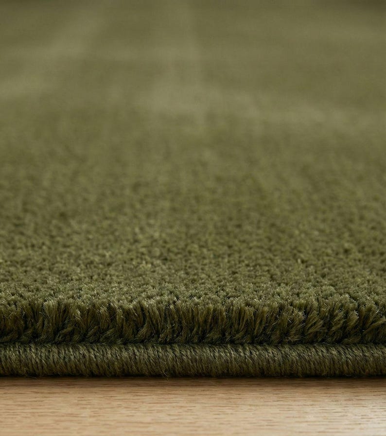 May include: Close-up of a plush, olive green rug. The rug has a soft, textured surface with a visible fringe along the edge. The color is a muted, earthy green, suggesting a natural or organic aesthetic. The rug appears to be made of a dense, high-pile material.