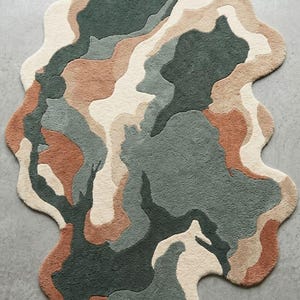 May include: An area rug with an irregular, organic shape featuring a tufted texture. The design consists of abstract, flowing color blocks in shades of dark forest green, muted sage, warm terracotta, and creamy off-white, creating a layered pattern.