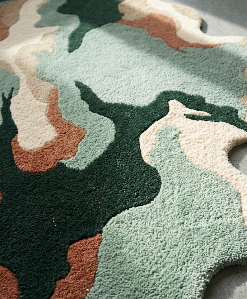 May include: An area rug with an irregular, organic shape featuring a tufted texture. The design consists of abstract, flowing color blocks in deep forest green, sage green, cream, and terracotta orange, creating a layered, topographical appearance.