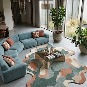 May include: An irregular, organic-shaped area rug featuring a multi-colored abstract pattern in shades of teal, terracotta, and cream. The rug is positioned on a concrete floor beneath a square glass coffee table with a light-toned wooden base.