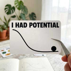 May include: A white sticker with the text "I HAD POTENTIAL" in black, featuring a black line graph with a ball at the bottom. The sticker is being held by tweezers, with plants and a notebook in the background.