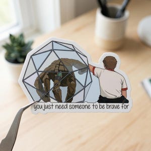 May include: A sticker featuring a cartoon illustration of a brown creature inside a geometric cage, with a person reaching out to touch it. The text "you just need someone to be brave for" is printed below the image.