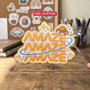 May include: A die-cut sticker featuring the word AMAZE repeated three times in orange lettering with blue outlines. A small orange geometric crab sits atop the text, surrounded by blue star accents and a blue orbital ring encircling the design.