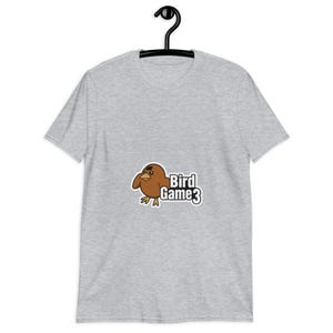 May include: A light gray t-shirt featuring a cartoon bird with a black eye mask and the text "Bird Game 3". The bird is brown with yellow feet and a yellow beak. The shirt is hanging on a black hanger.