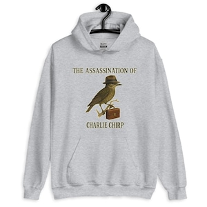 May include: A light gray hooded sweatshirt with the text "THE ASSASSINATION OF CHARLIE CHIRP." The graphic features a bird wearing a hat and holding a briefcase. The bird is perched on a branch.