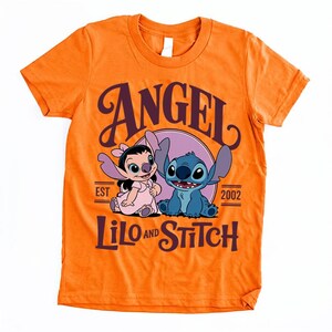 May include: Bright orange t-shirt with a cartoon design of Angel and Stitch from Lilo and Stitch. The design includes the words "ANGEL" and "LILO AND STITCH" in a dark purple colour. The shirt also has the text "EST 2002".