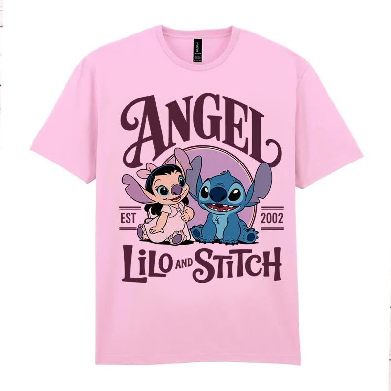 May include: A pale pink t-shirt with a graphic of Angel and Stitch from Lilo & Stitch. The design includes the word "ANGEL" in a decorative font above the characters, with "LILO AND STITCH" below. The text "EST 2002" is also included.