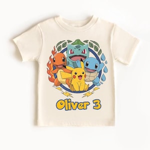 May include: Cream-colored t-shirt featuring a colorful graphic of four cartoon characters inside a blue and green circular design. The text "Oliver 3" is printed in yellow and blue below the graphic. The shirt is made of soft material.