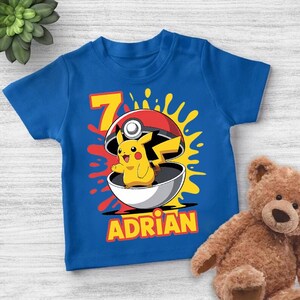 Pode incluir: Camiseta azul com um desenho de Pikachu saindo de uma Poké Bola, com o número 7 e o nome "ADRIAN" em amarelo. O design inclui respingos vermelhos e amarelos. Um ursinho de pelúcia está no fundo.