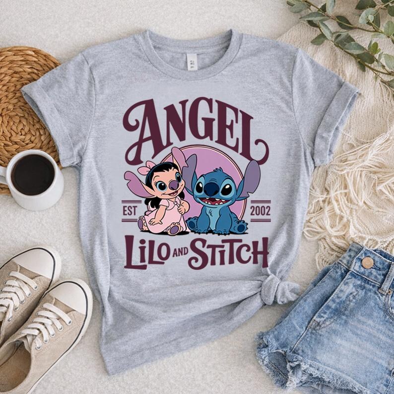 May include: A light grey t-shirt with a design featuring Angel and Stitch from Lilo & Stitch. The design includes the word "ANGEL" in burgundy script above the characters, with "LILO and STITCH" below. The shirt has rolled-up sleeves.