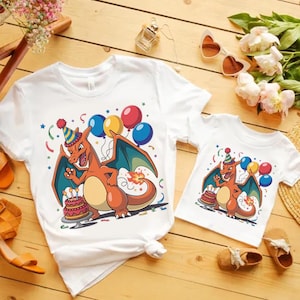May include: Two white t-shirts featuring a cartoon Charizard design, celebrating a birthday with a cake, balloons, and confetti. One shirt is adult size, the other is child size. The shirts are on a wooden surface.