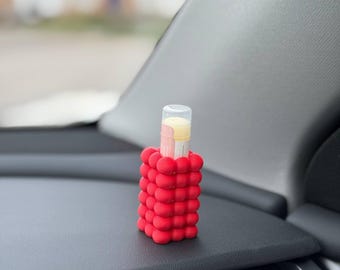 May include: A red, bubble-textured holder encases a clear tube containing a lip balm. The lip balm has a pink and yellow color scheme. The holder is sitting on a black surface.