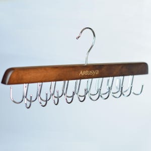 May include: A wooden tie rack with a metal hook and multiple metal hooks for hanging ties. The rack is made of dark brown wood and has the word "Artusya" printed on it.
