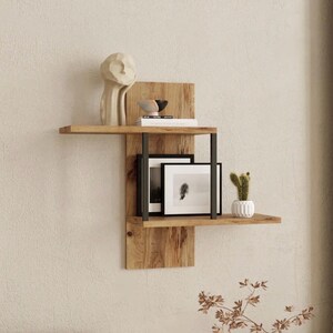 May include: A two-tiered wooden wall shelf with black metal supports. The shelf displays a cream-colored sculpture, small decorative bowls, books, framed black and white photos, and a small potted cactus. The wood has a natural grain.