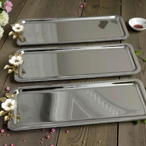 Op de afbeelding: Drie rechthoekige serveerschalen van roestvrij staal. Elke schaal heeft een decoratief bloemaccent met witte bloemen en goudkleurige bladeren. De schalen zijn gerangschikt op een houten oppervlak.
