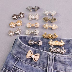 Pode incluir: Uma variedade de botões e fechos decorativos para jeans em vários designs, incluindo formas florais, de laço e geométricas. Os botões são em tons de prata, ouro e bronze, alguns com detalhes de pérolas. Os botões estão presos a uma cintura de jeans de ganga.