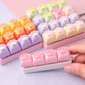 Könnte beinhalten: Eine Sammlung farbenfroher Tastenkappen mit Pfotenabdruck-Designs. Die Keycaps sind in verschiedenen Pastellfarben gehalten, darunter Gelb, Orange, Rosa, Lila und Grün. Die Pfotenabdrücke sind in kontrastierenden Farben gehalten.
