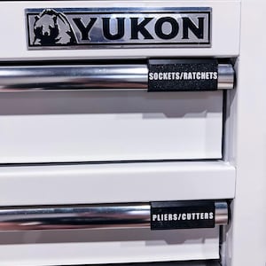 May include: A white tool chest with chrome drawer pulls. The top drawer label reads "SOCKETS/RATCHETS" and the bottom drawer reads "PLIERS/CUTTERS". The chest has a "YUKON" logo.