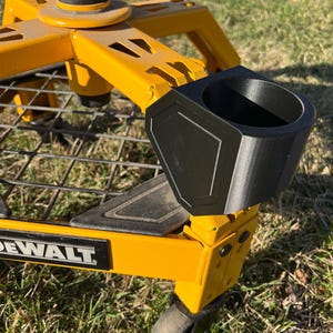 May include: A black cup holder attached to a yellow metal cart. The cup holder has a geometric design and is mounted on the side of the cart. The cart has the word "DEWALT" printed on it. The cart is on a grassy surface.